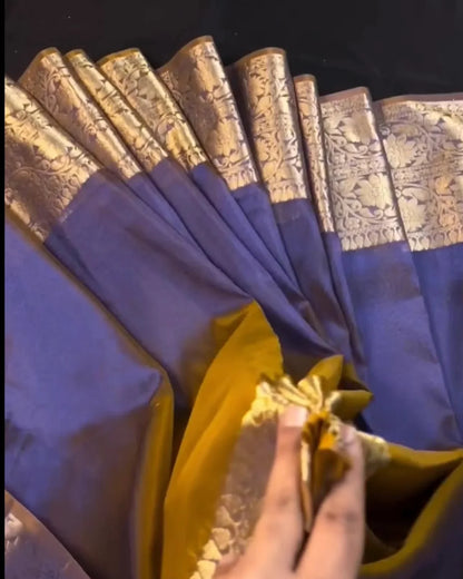 A close-up of a purple and gold saree being held open by a hand, showing its metallic zari border and yellow-gold inner layer against a dark background.