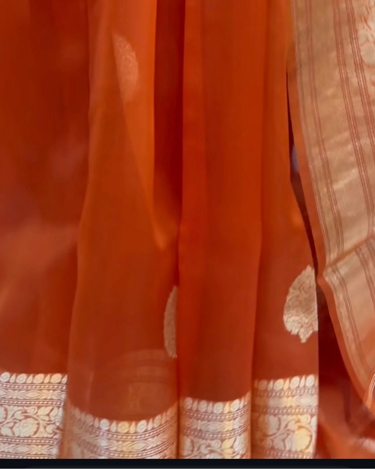 close-up of an orange saree fabric with a gold and silver patterned border and a matching orange and gold patterned drape.