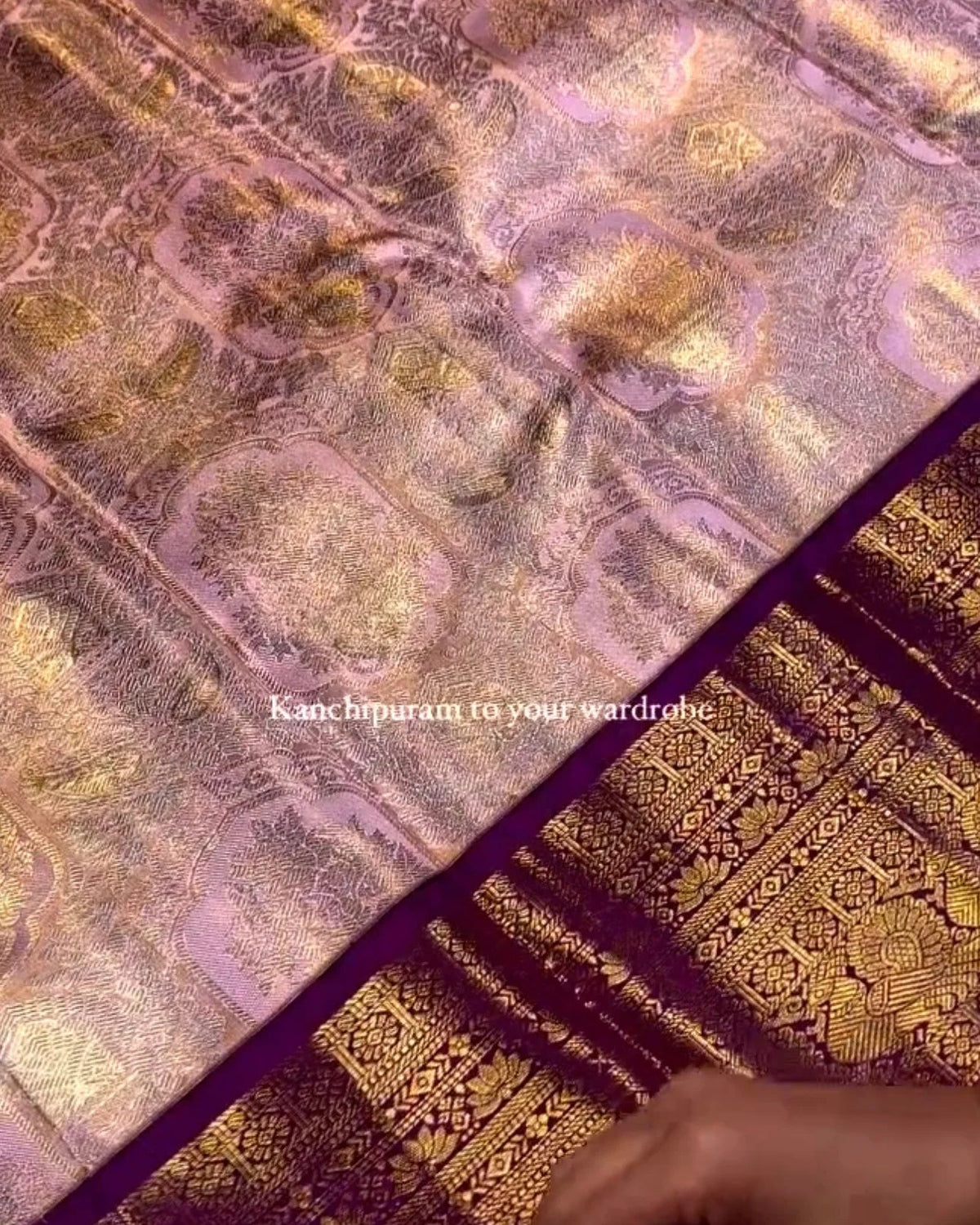 A close-up of a purple and gold patterned fabric, likely a saree or textile, with ornate metallic gold motifs and a deep purple border. A person's hands are visible at the bottom right, holding the fabric.