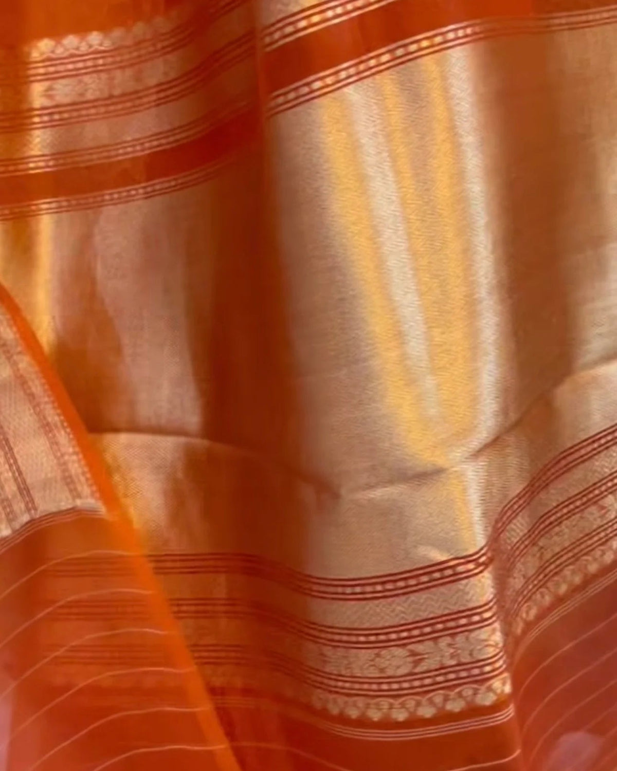 Close-up of an orange and gold woven fabric with metallic zari stripes and ornate border patterns.