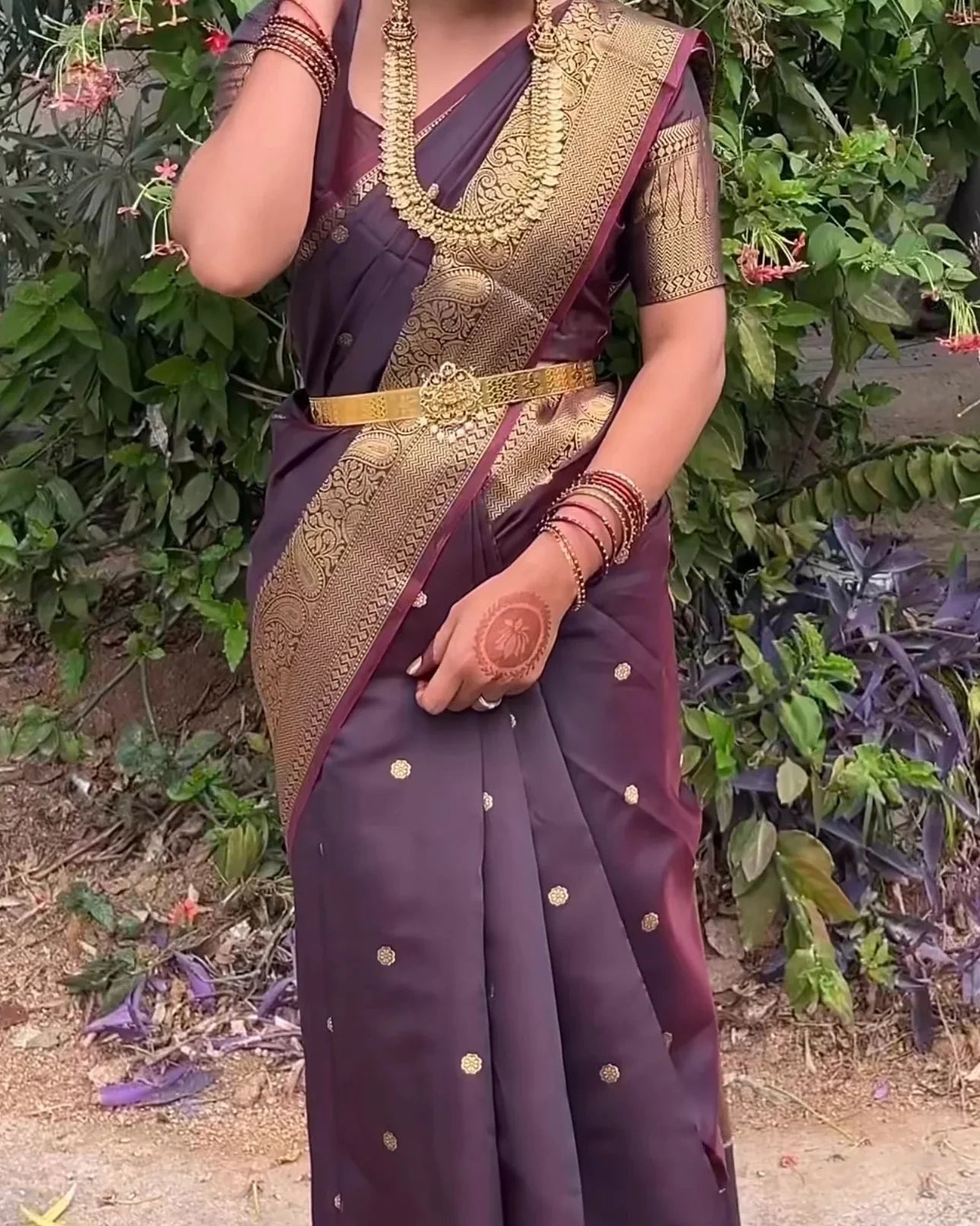 a woman wearing a dark purple saree with a wide gold border, gold zari motifs, and a gold belt, standing outdoors in front of green foliage.