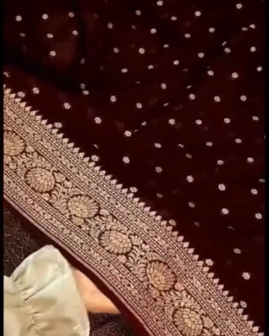 A close-up of a dark maroon or burgundy fabric with small white polka dots and a wide ornate beige embroidered border. A small portion of a light-colored sleeve is visible at the lower left.