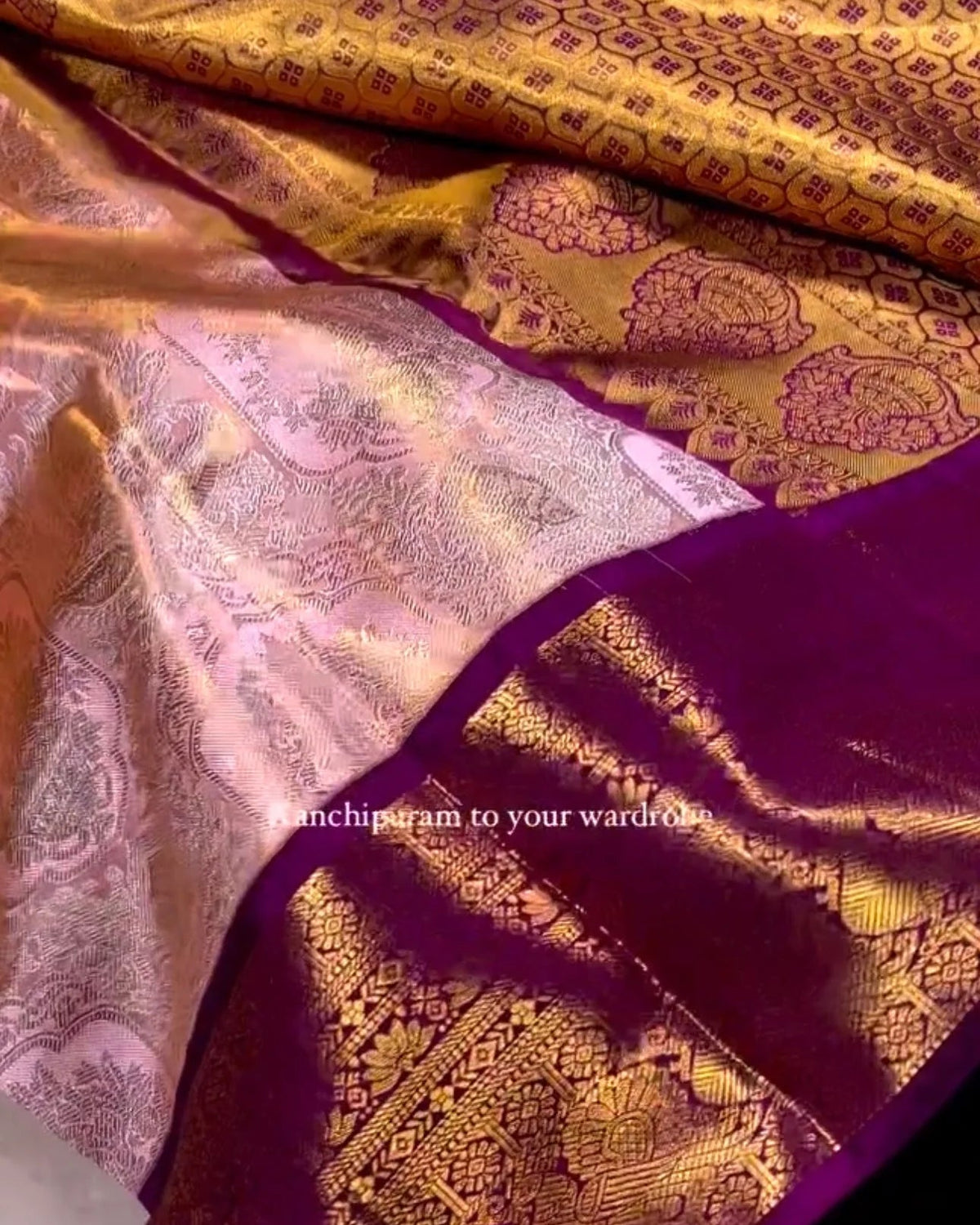 A close-up of a luxurious gold and pink Banarasi silk saree with a deep purple border, held by a hand to show the fabric's texture and sheen.