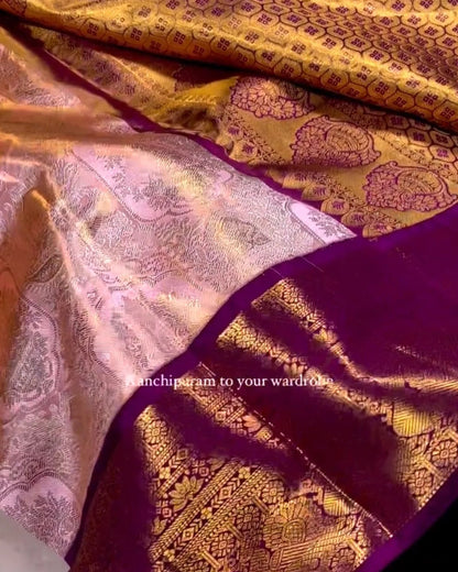 A close-up of a luxurious gold and pink Banarasi silk saree with a deep purple border, held by a hand to show the fabric's texture and sheen.