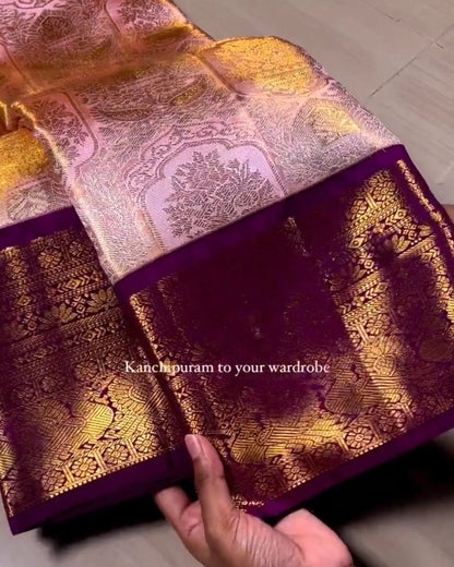 A close-up of a hand holding up a purple and gold brocade fabric, showing its intricate woven patterns and border. The background is a light-colored wooden floor.