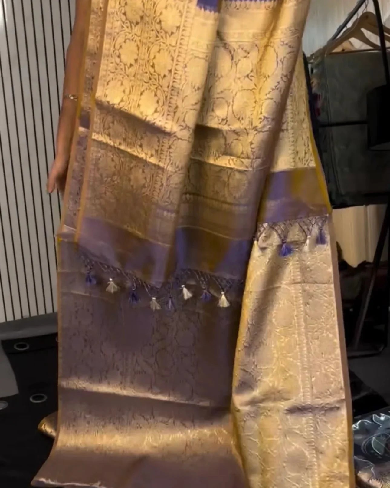 a close-up of a draped silk saree with a metallic gold body, intricate woven patterns, and a lavender-purple border with tassel details.