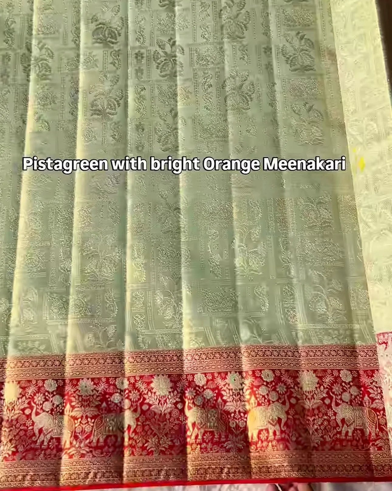 a folded and pleated light pistachio green banarasi silk saree with intricate gold zari floral motifs, featuring a prominent orange and red floral border.