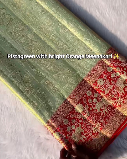 A folded pistachreen silk saree with a bright orange and gold Meenakari border, held by a hand on a white textured background.