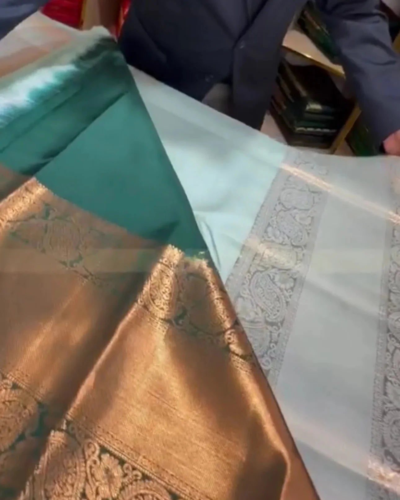 A close-up of a traditional Indian silk saree, showing a teal green body, a wide metallic gold zari border, and a white border with intricate silver paisley motifs.
