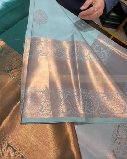 A light blue-green silk saree with a wide metallic gold and silver brocade border, being draped over a table. A person's hands and torso are visible holding the fabric.