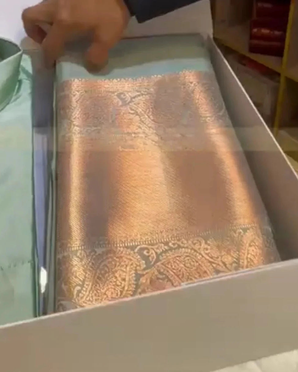 A folded metallic gold and silver patterned silk scarf or sash is displayed inside a white box next to a light green shirt, with a hand holding the top edge of the fabric.