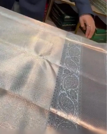 A close-up of a metallic silver fabric, likely a saree, showing its woven texture, sheen, and a detailed paisley border.