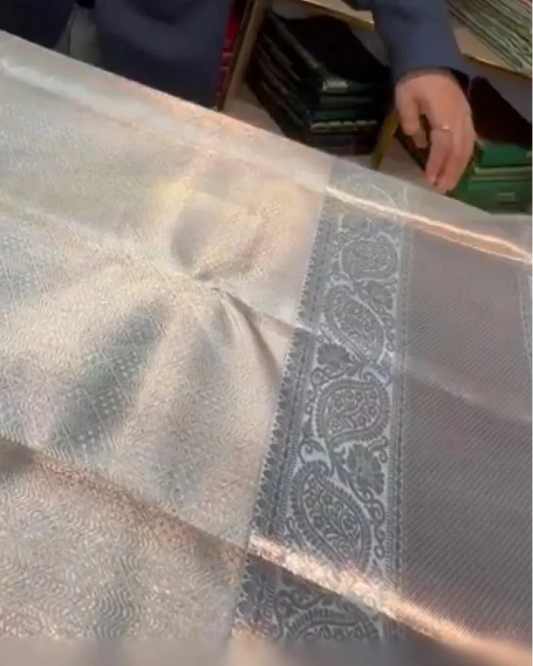 A close-up of a metallic silver fabric, likely a saree, showing its woven texture, sheen, and a detailed paisley border.