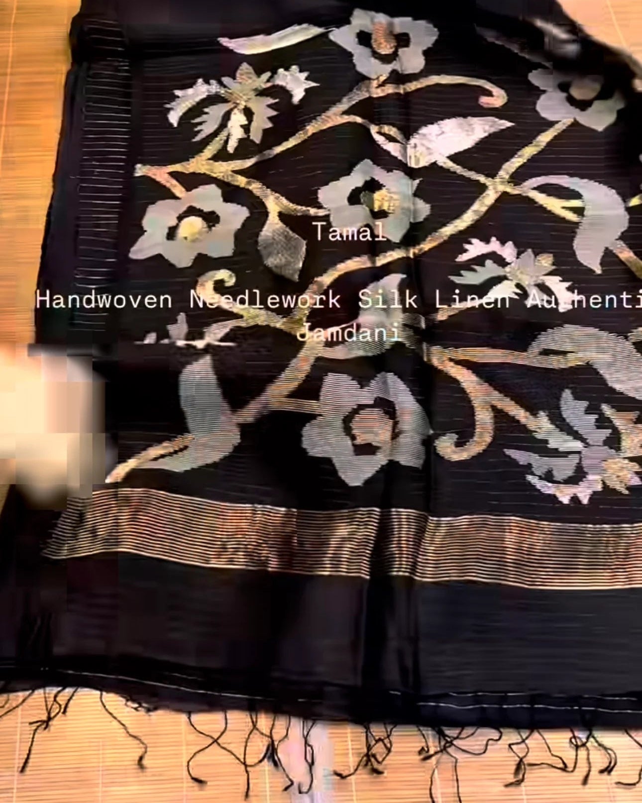 a black handwoven silk linen augmenti damask textile with intricate floral embroidery in gold, silver, and gray, laid flat on a light-colored wooden surface.