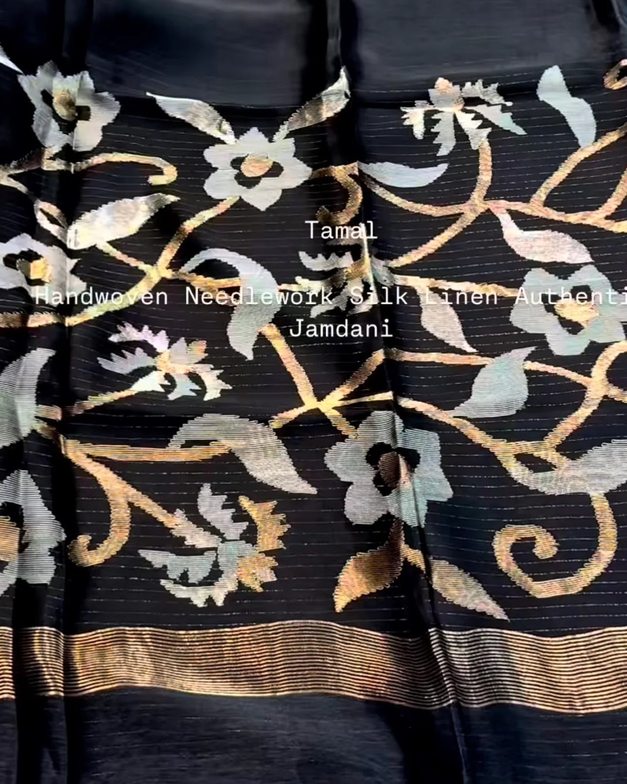black silk saree fabric with intricate jamdani floral embroidery in gold, beige, and light blue, featuring a decorative gold border and fringed edges.
