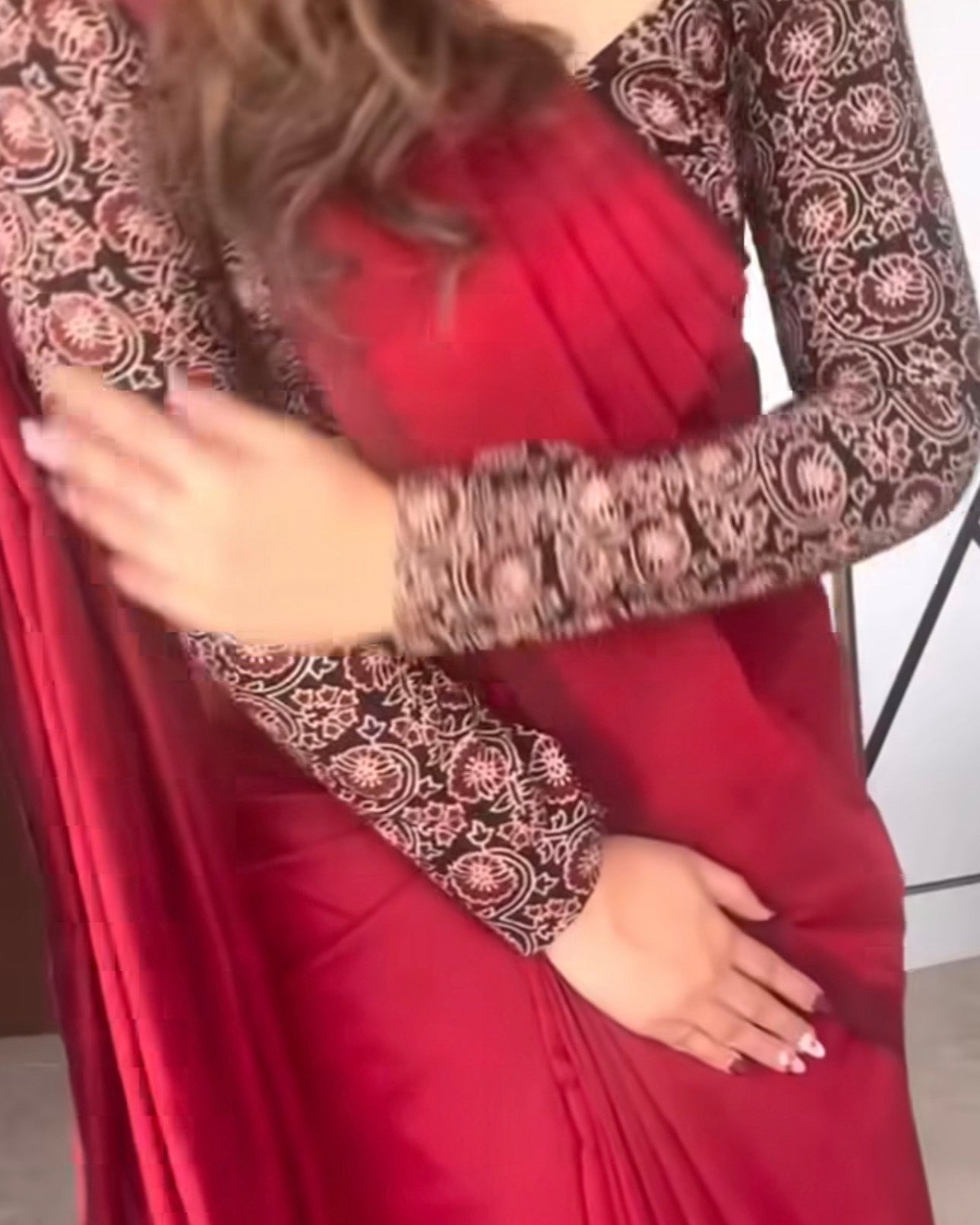 a woman wearing a red saree with long sleeves featuring intricate black and beige floral embroidery, posed indoors.