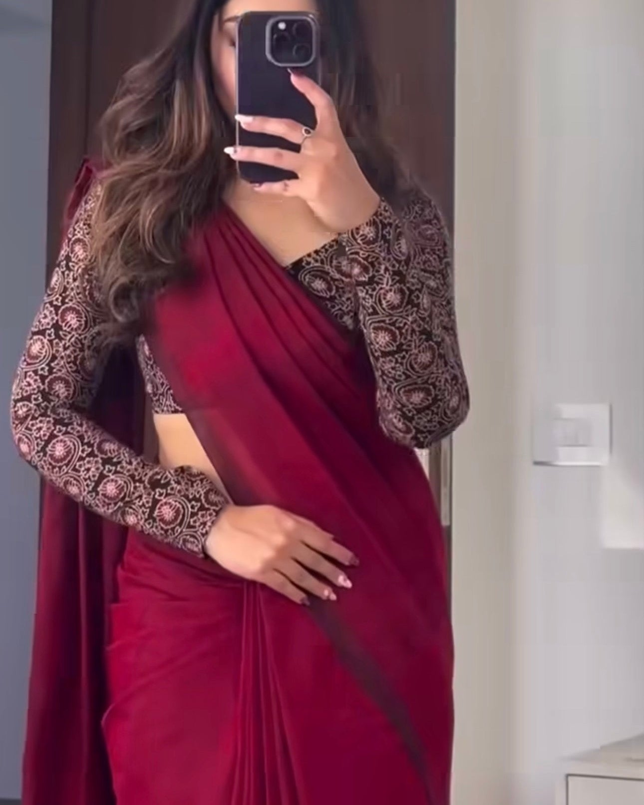 A woman takes a mirror selfie wearing a deep red sari with a black and gold patterned long-sleeve blouse in an indoor setting.
