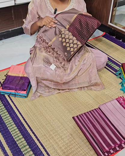 A woman sitting on a woven mat displays a folded purple and gold patterned fabric, with other folded fabrics stacked nearby on the floor.
