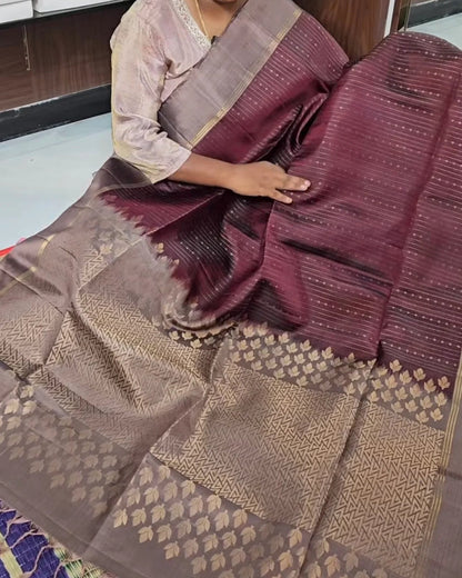 A woman seated indoors wearing and displaying a dark plum purple and brown silk saree with metallic gold zari woven patterns and borders.