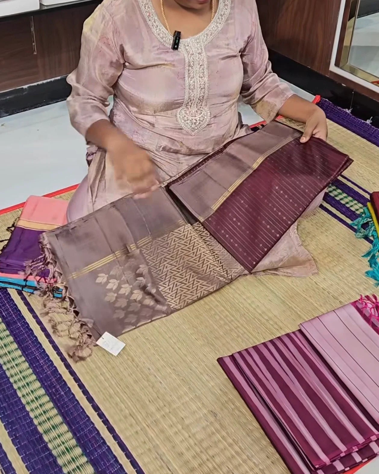 A person seated on a woven mat holds up a folded piece of purple and mauve fabric with metallic gold woven patterns and tassels, surrounded by other folded textile pieces in a studio or workshop setting.