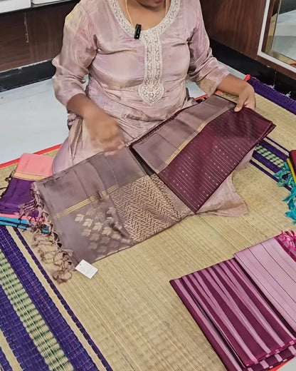 A person seated on a woven mat holds up a folded piece of purple and mauve fabric with metallic gold woven patterns and tassels, surrounded by other folded textile pieces in a studio or workshop setting.